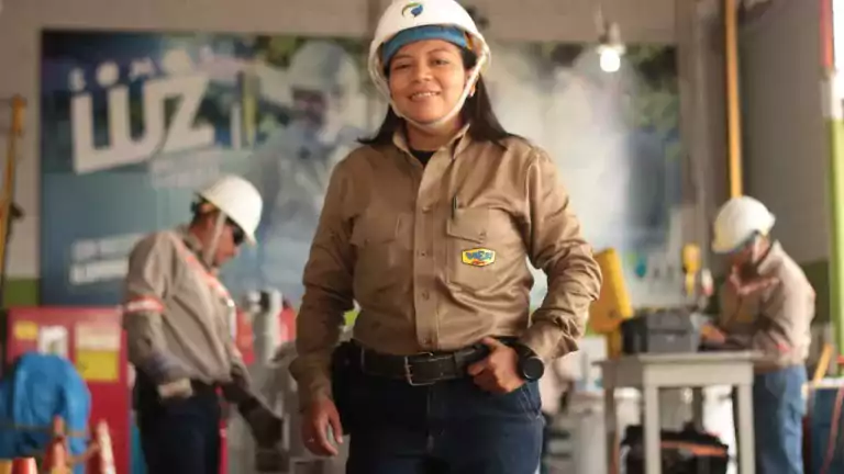 Woman wearing a hard hat and work uniform stands confidently in an industrial setting with two coworkers in the background, both also wearing safety gear. The environment appears to be a workshop or factory, emphasizing workplace safety and teamwork.