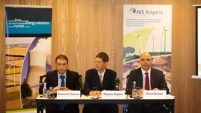 Three men in business attire sit at a conference table with microphones and nameplates in front of AES Bulgaria banners promoting sustainable energy solutions, with images of wind turbines and a power plant in the background.