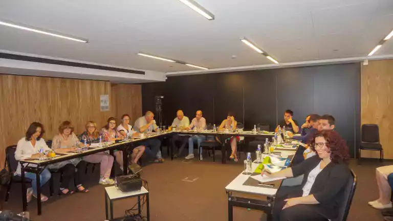 A group of people seated around a U-shaped conference table in a modern meeting room, engaged in discussion with notebooks, water bottles, and papers in front of them. The room has wooden panel walls and bright ceiling lights.