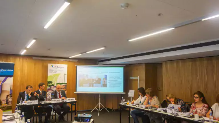 A group of people in a conference room attending a business meeting. Three men in suits sit at a table with nameplates, while several women sit on the right side taking notes. A projector displays a presentation on a screen. Banners and water bottles are visible on the tables.