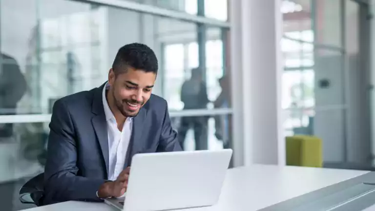 Smiling man in a business suit working on a laptop at a modern office desk with glass walls in the background.