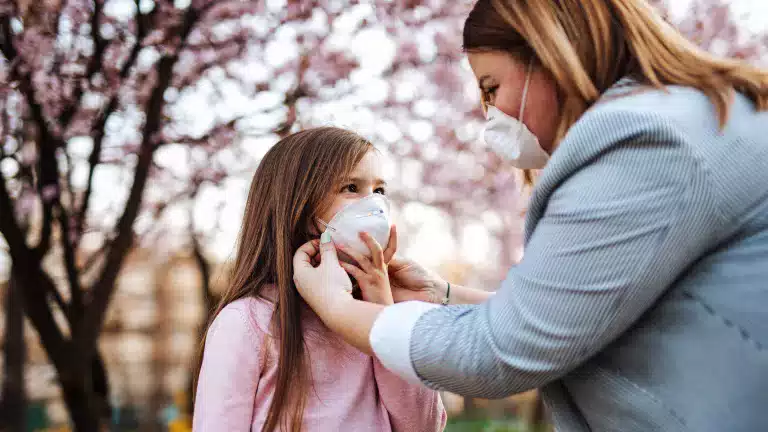 An adult helps a young girl put on a protective face mask outdoors, with blooming pink trees in the background. Both are wearing masks, and the scene suggests care and safety during springtime.