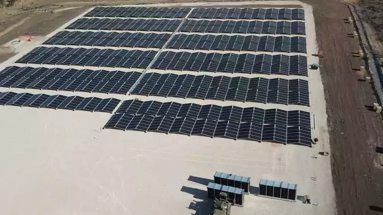 Aerial view of a large solar farm with multiple rows of solar panels installed on a cleared, sandy area surrounded by trees and dirt roads. Equipment and containers are visible near the bottom of the image.