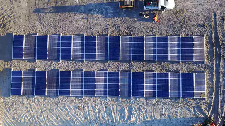 Aerial view of a solar panel installation site with two rows of blue solar panels on sandy ground. Construction vehicles, workers, and equipment are visible near the panels, indicating ongoing work.