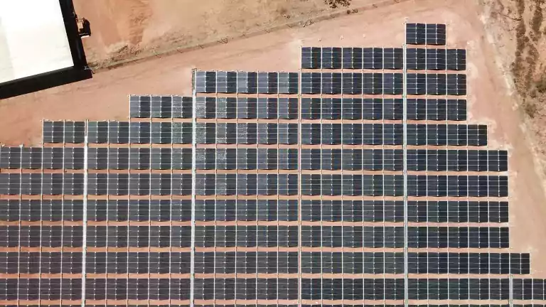 Aerial view of a large solar panel array installed on reddish soil in a remote, arid area. The solar panels are arranged in neat rows and columns, surrounded by sparse vegetation and industrial equipment nearby.
