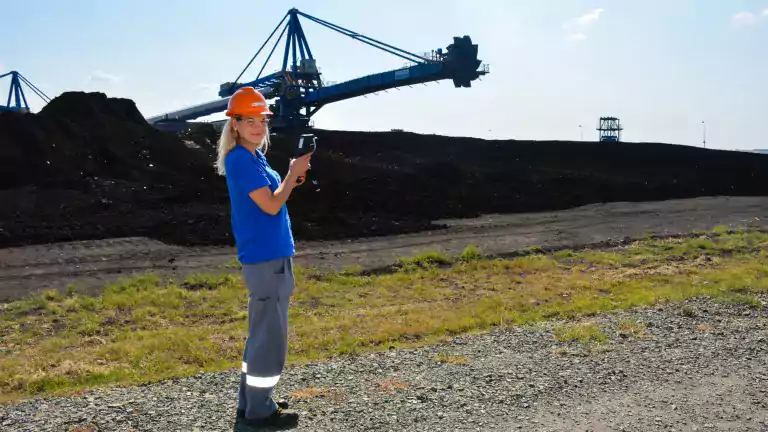 A woman wearing a hard hat and safety glasses stands on a gravel path at a mining site, holding a handheld device. Behind her are large piles of coal and a blue industrial excavator under a clear sky.