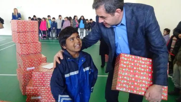 A man in a blue jacket gives a wrapped gift box to a smiling young boy in a gymnasium, with other children and stacked gift boxes visible in the background.