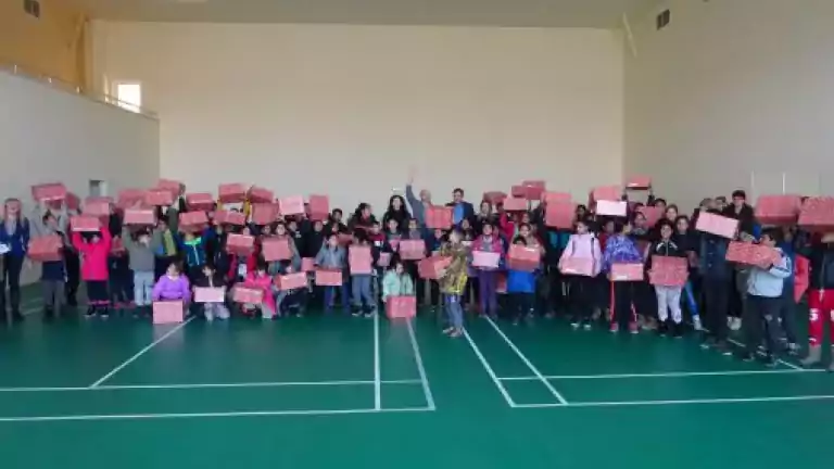 Large group of children and adults standing in a gymnasium holding red and white gift boxes, posing for a group photo on a green sports court.