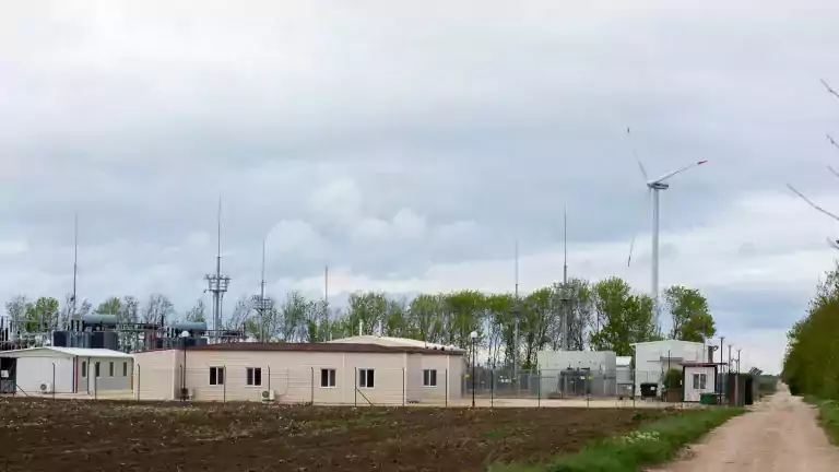 A rural wind farm facility with several small buildings, antennas, and a large wind turbine in the background, surrounded by trees and a dirt road under a cloudy sky.