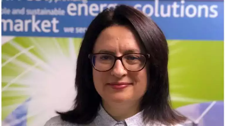 Woman with dark hair and glasses wearing a white shirt standing in front of a colorful background with text about energy solutions and a wind turbine image.