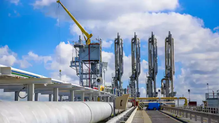 Industrial oil and gas facility with large pipelines, metal structures, and a yellow crane under a partly cloudy sky. The scene shows a modern energy infrastructure with equipment for handling or transporting fuel.