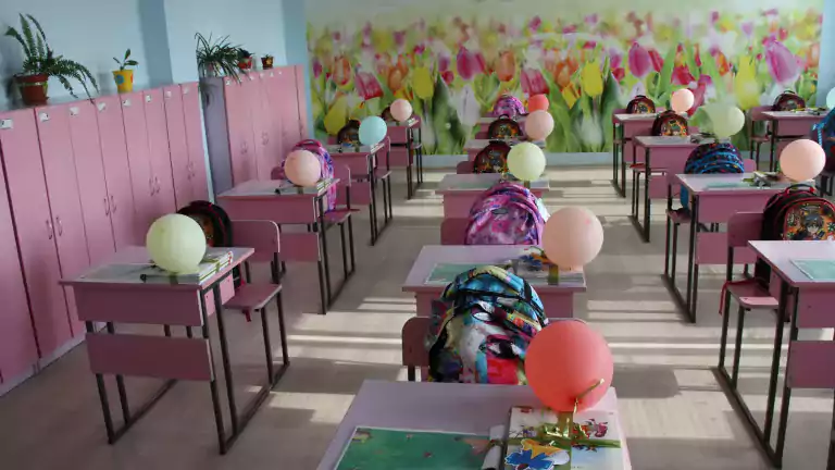 A bright, colorful classroom with pink desks and chairs, each desk decorated with a balloon, books, and a backpack. The classroom has pink lockers, potted plants on top, and a wall mural of tulips in the background.