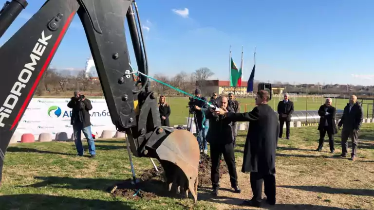 A group of people participates in a groundbreaking ceremony outdoors, with a large excavator and a man cutting a green ribbon. Photographers and onlookers stand nearby, while Bulgarian flags and a banner are visible in the background under a clear blue sky.