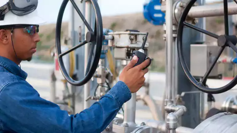 Industrial worker wearing safety gear and a hard hat operates large valve controls at an outdoor facility, using a handheld radio for communication.