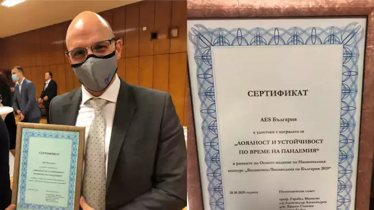 A man in a suit and face mask holds a framed certificate at an indoor event. The certificate, written in Bulgarian, recognizes AES Bulgaria for loyalty and resilience during the pandemic. Other attendees in formal attire and masks are visible in the background.
