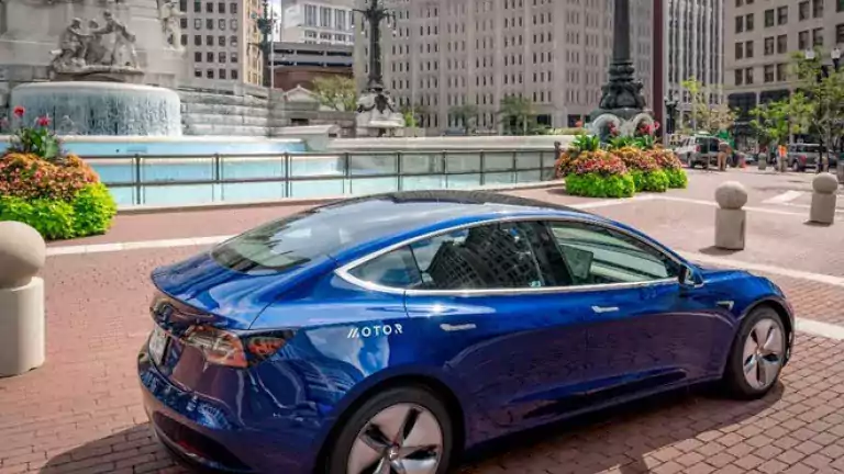 A blue electric car parked on a brick plaza near a large city fountain, surrounded by tall buildings and decorative flower beds on a sunny day.