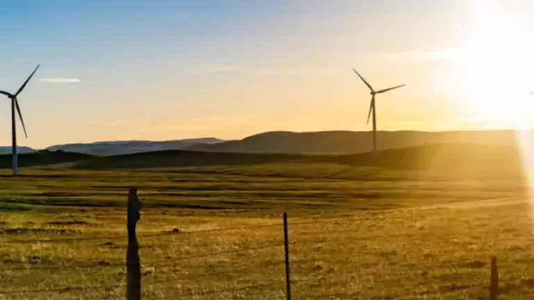 Wind turbines on a grassy field with a wooden fence in the foreground and the sun setting over distant hills, under a clear blue sky.