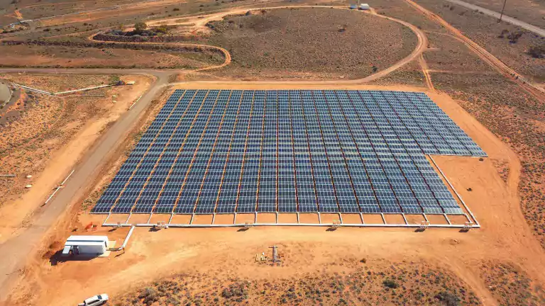 Aerial view of a large solar farm with rows of solar panels installed on a flat, dry landscape surrounded by dirt roads and sparse vegetation.