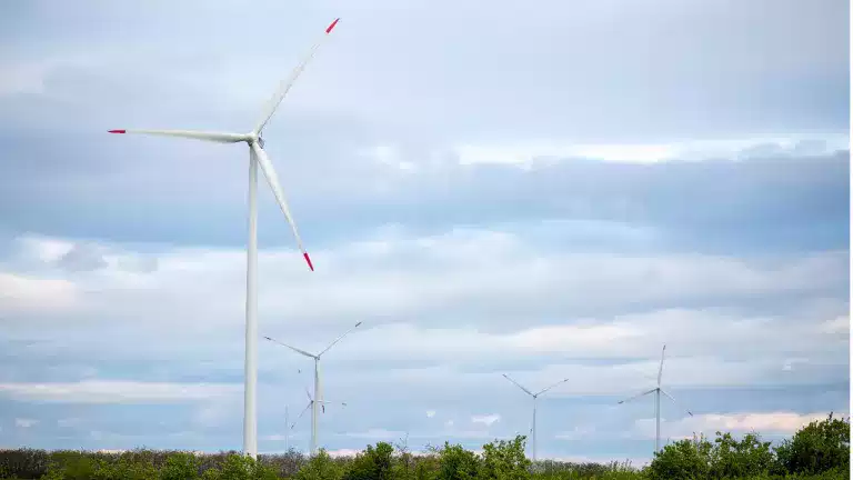 Several wind turbines stand in a green field under a cloudy sky, generating renewable energy. The largest wind turbine is in the foreground, with others visible in the background among trees and grass.