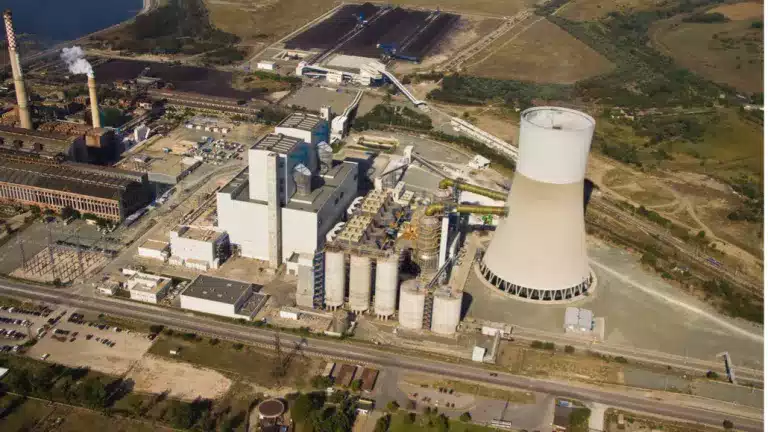 Aerial view of a large industrial power plant with a prominent cooling tower, multiple silos, and surrounding buildings, set in a rural landscape with roads and greenery.