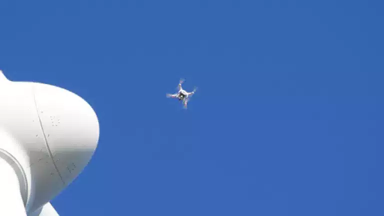 A close-up of a white wind turbine with a small drone flying nearby against a clear blue sky.