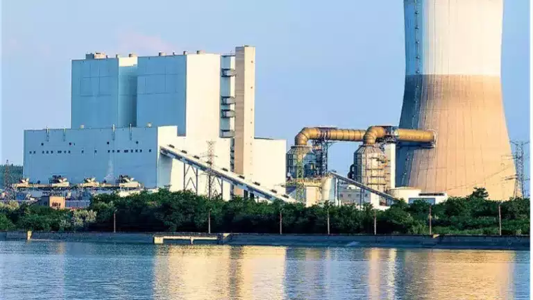 A large industrial power plant with a cooling tower and several buildings is situated near a body of water, surrounded by greenery under a clear sky.