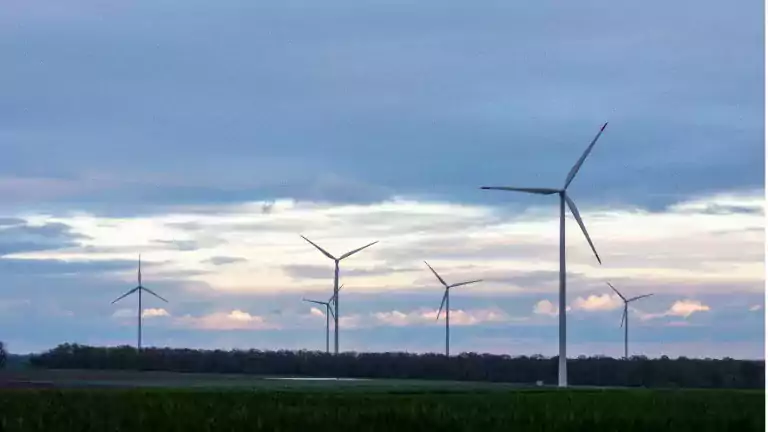 Several wind turbines stand in a green field under a cloudy sky at sunset, generating renewable energy in a rural landscape.