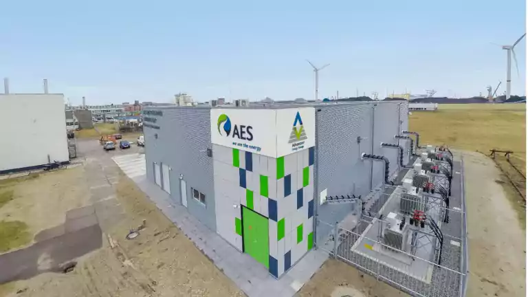 Modern energy facility with AES and Advancer logos on the building, surrounded by fenced electrical equipment and wind turbines in the background, under a clear blue sky.