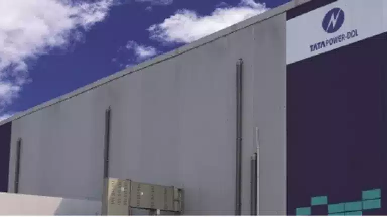 Modern industrial building with logos of Tata Power-DDL, AES, Mitsubishi Corporation, and Fluence on the exterior wall, set against a partly cloudy blue sky.