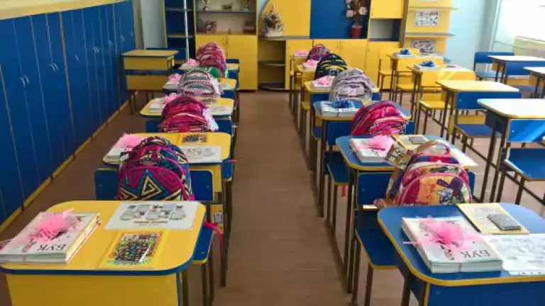 Colorful classroom with rows of yellow and blue desks, each desk set with books, pink decorations, and patterned backpacks. The classroom is tidy and well-organized, with cabinets and shelves along the walls and sunlight coming through the window.