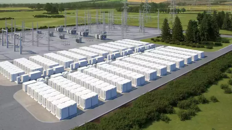 Aerial view of a large battery energy storage facility with rows of white battery containers and electrical substation equipment, surrounded by greenery and trees.