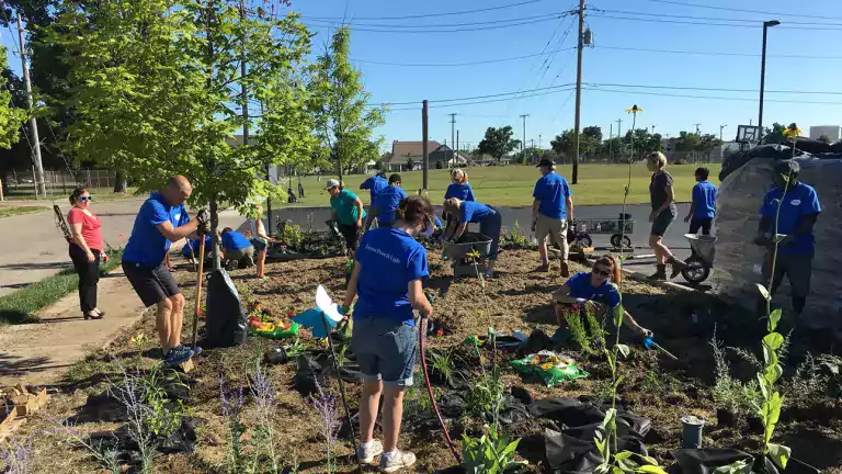 A group of volunteers in blue shirts plant trees and flowers in a community garden on a sunny day. Some people use shovels, others water plants, and a few supervise. The setting is a neighborhood with open green space and utility poles in the background.