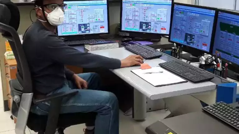 A person wearing a white hard hat, safety goggles, and a mask sits at a desk in a control room, monitoring four computer screens displaying technical data and system controls.