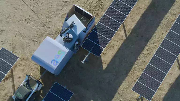 Aerial view of a robotic machine inspecting or cleaning solar panels in a solar farm on dry ground. The robot is positioned between rows of solar panels and appears to be designed for solar panel maintenance.