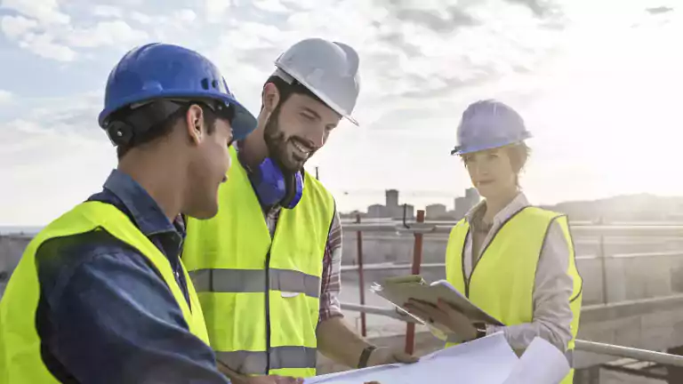 Three construction workers wearing safety vests and helmets review blueprints and documents at a building site under a cloudy sky.