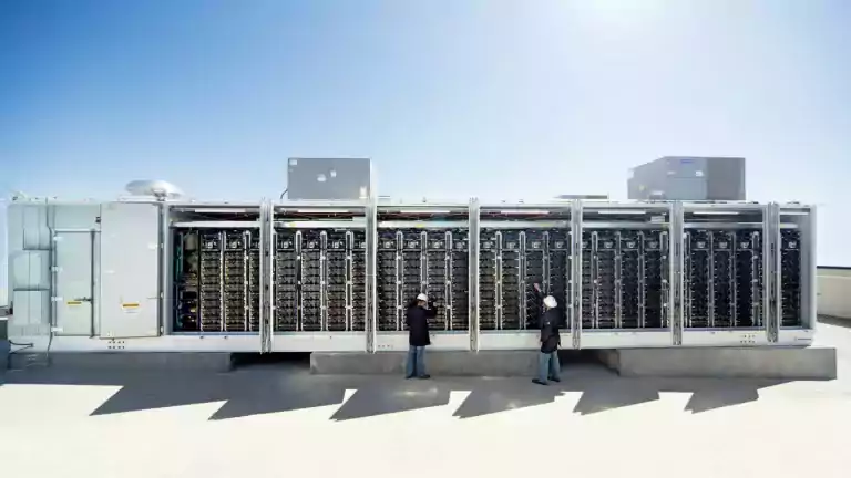 Two engineers wearing hard hats inspect a large outdoor data center server unit on a rooftop under a clear blue sky. The server racks are exposed, showing rows of computer hardware and cooling systems.
