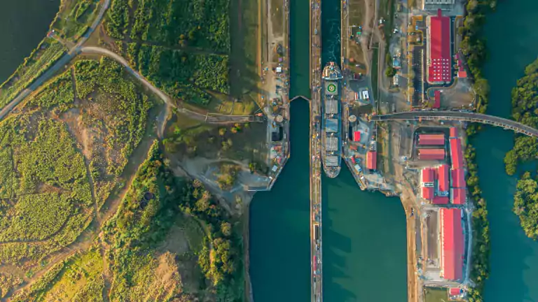 Aerial view of a large ship passing through a canal lock system surrounded by green vegetation, roads, and red-roofed buildings on both sides of the waterway.