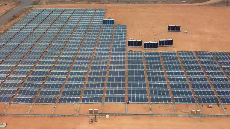 Aerial view of a large solar farm with rows of solar panels installed on a dry, sandy landscape, surrounded by desert terrain and a few scattered structures.