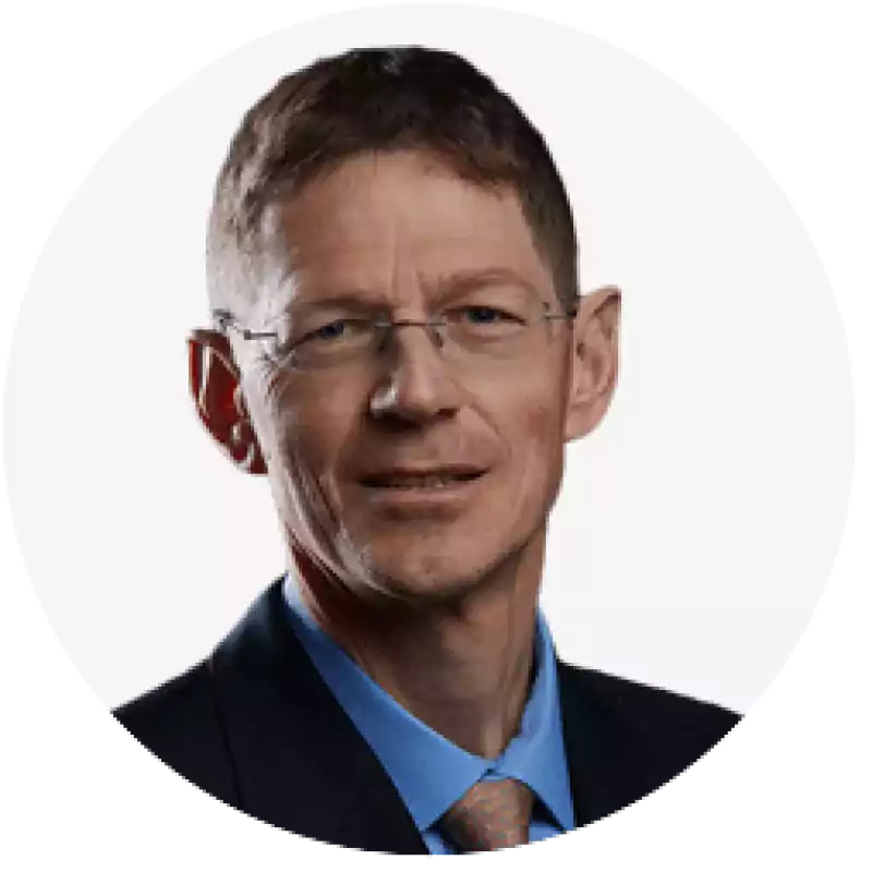 Portrait of a middle-aged man with short brown hair, wearing glasses, a dark suit, blue shirt, and patterned tie, against a plain white background.