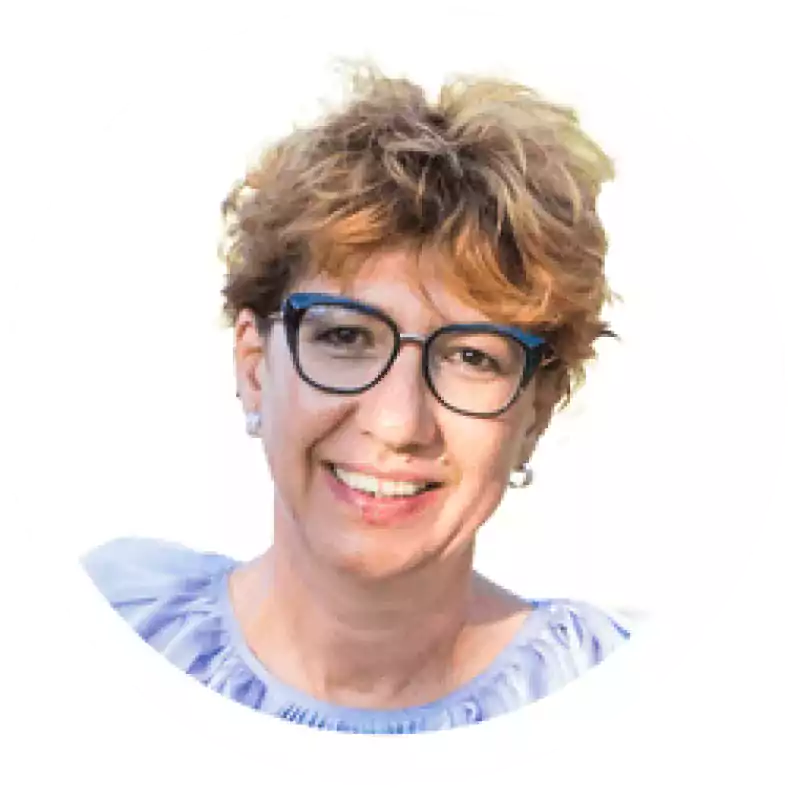 Smiling middle-aged woman with short curly hair and glasses wearing a light blue blouse, photographed against a white background.