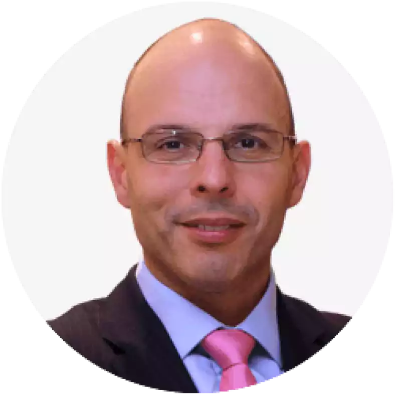 Portrait of a bald man wearing glasses, a dark suit, light blue shirt, and pink tie, smiling at the camera against a plain white background.
