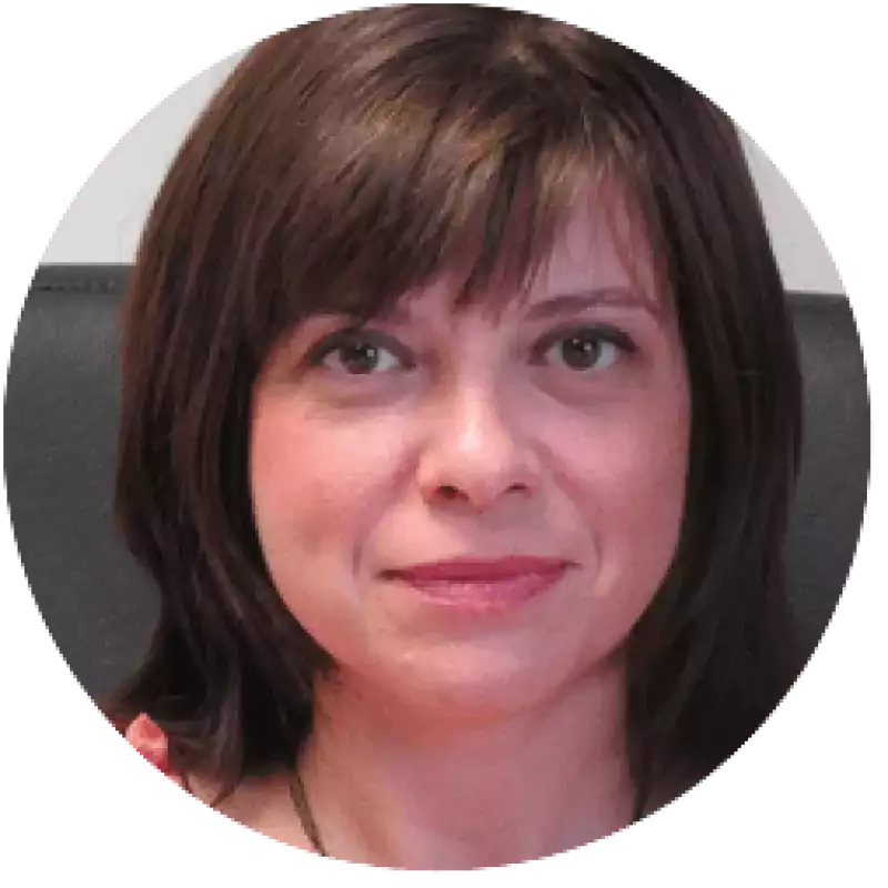 Portrait of a woman with straight brown hair, light skin, and brown eyes, sitting in front of a gray background, looking directly at the camera with a neutral expression.