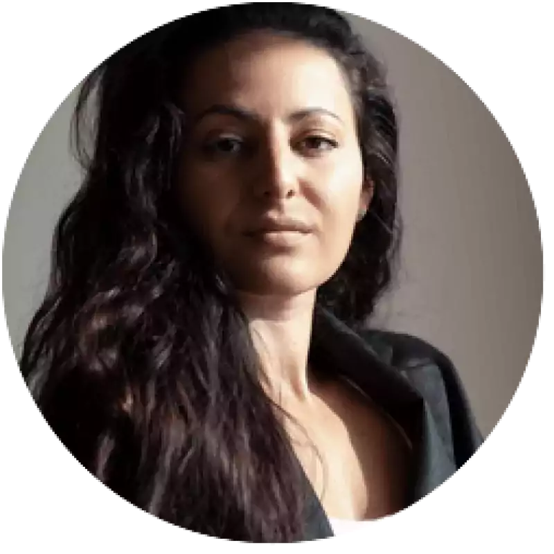 Woman with long wavy dark hair wearing a dark blazer, looking confidently at the camera in natural light with a neutral background.