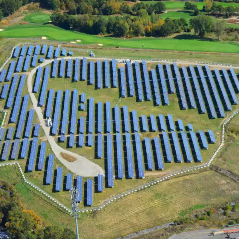 Aerial view of a large solar farm with rows of blue solar panels arranged on green grass, surrounded by trees and a golf course in the background.