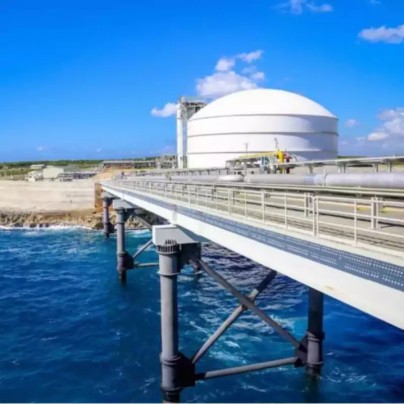 A large white liquefied natural gas storage tank is situated near the coastline, with a metal pipeline bridge extending over blue ocean water under a clear sky.