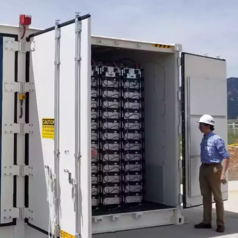 A person wearing a hard hat stands beside an open container filled with industrial batteries, likely part of an energy storage system, outdoors on a sunny day.