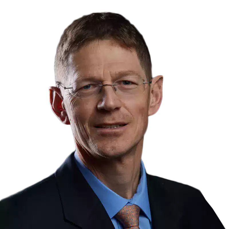 Portrait of a middle-aged man with short brown hair, wearing glasses, a dark suit, blue shirt, and patterned tie, against a dark gradient background.