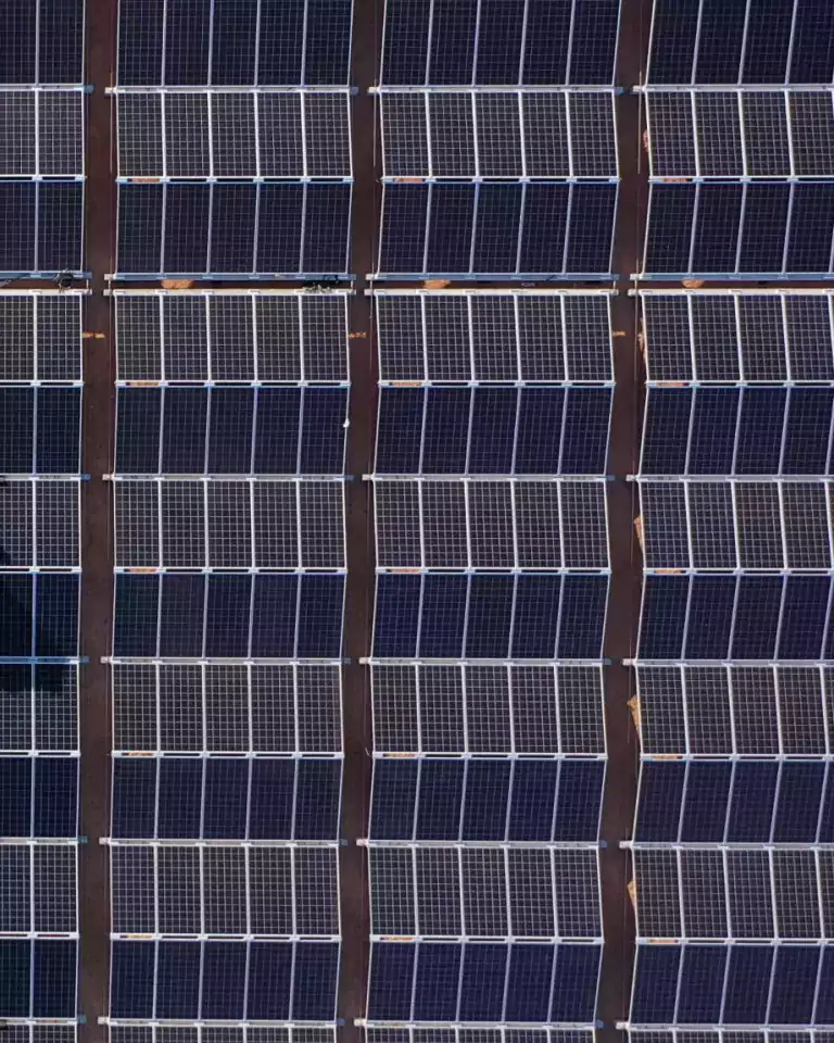 Aerial view of a tractor installing solar panels on a large solar farm in a desert area, with rows of solar panels aligned next to bare soil.