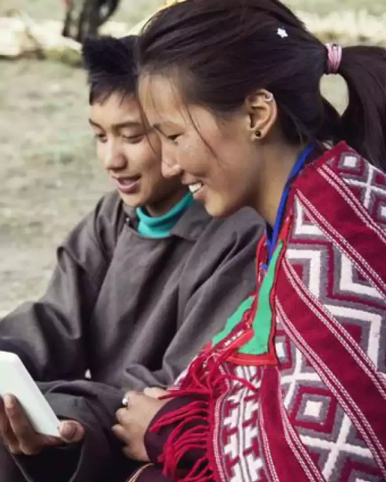 Two people sitting outdoors, smiling and looking at a tablet together. One person is wearing a traditional patterned red shawl, and both appear engaged and happy.