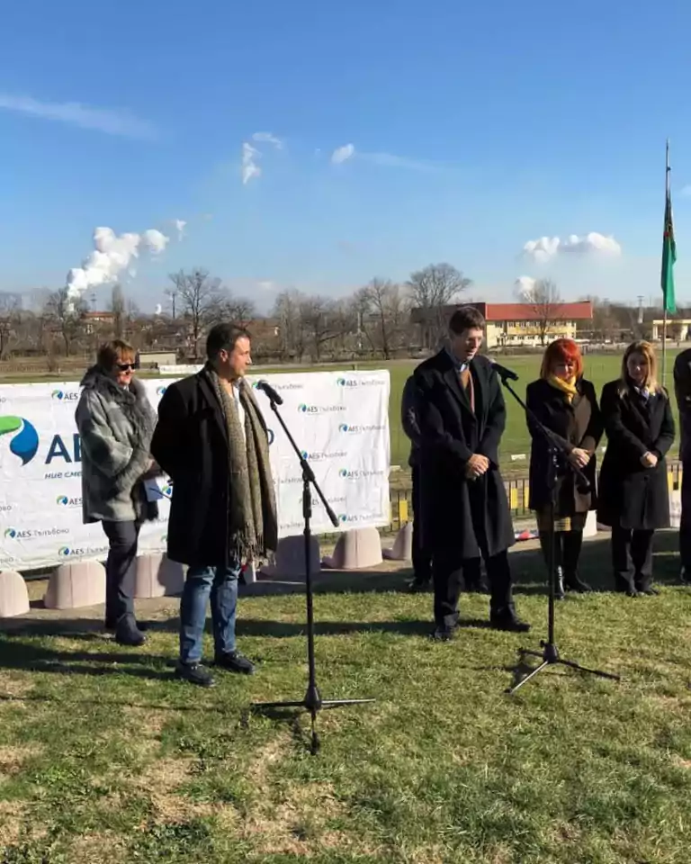 A group of people stands outdoors on grass, speaking at microphones during a formal event. A branded banner and flag are visible in the background, with clear blue sky and some smoke rising from a distant building. The participants are dressed in winter clothing.